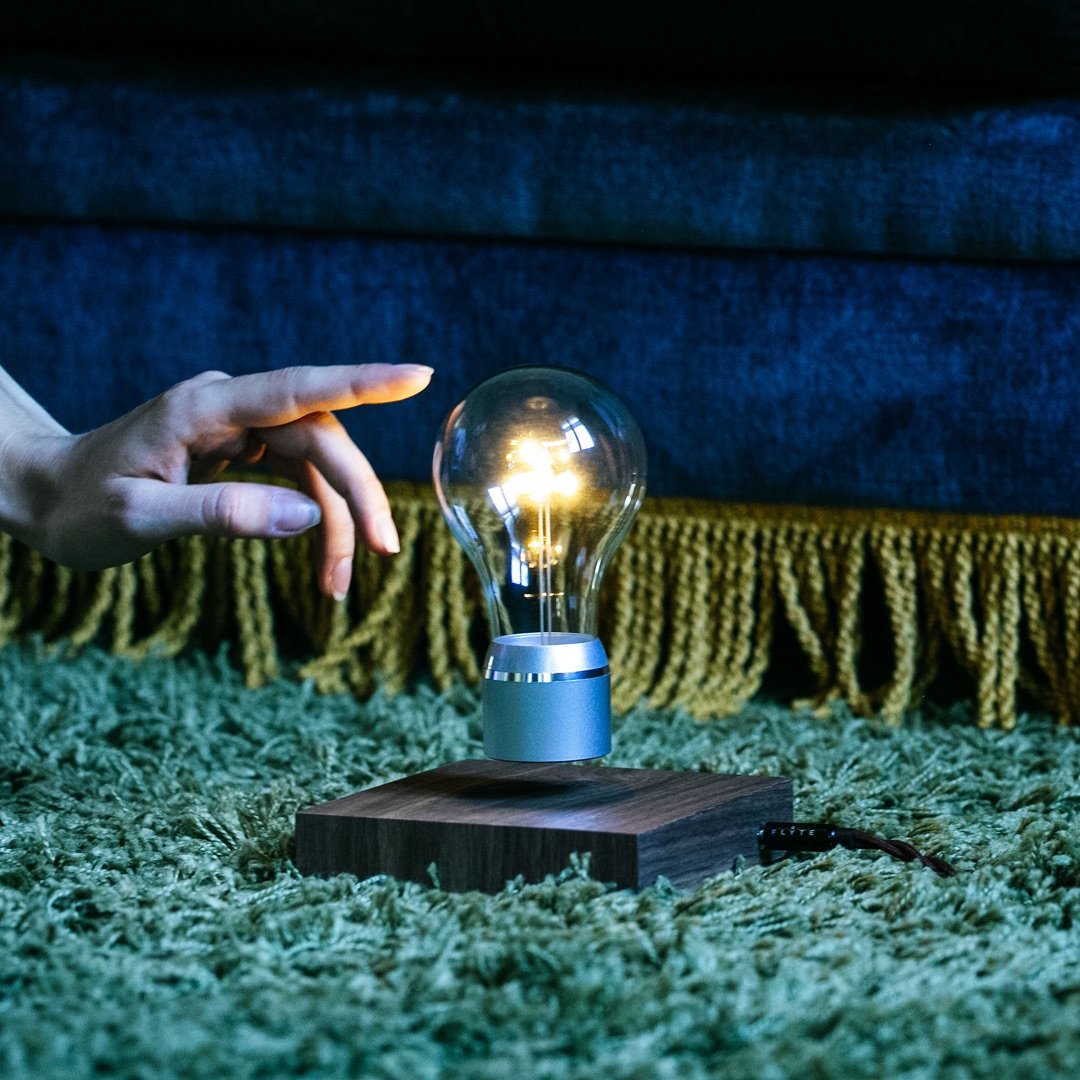 Levitating light bulb Light by Flyte, chrome cap bulb and walnut base version, placed on a carpet showing a hand reaching to the bulb